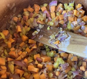 Wooden spatula stirring onion, garlic, sweet potato and banana peppers.